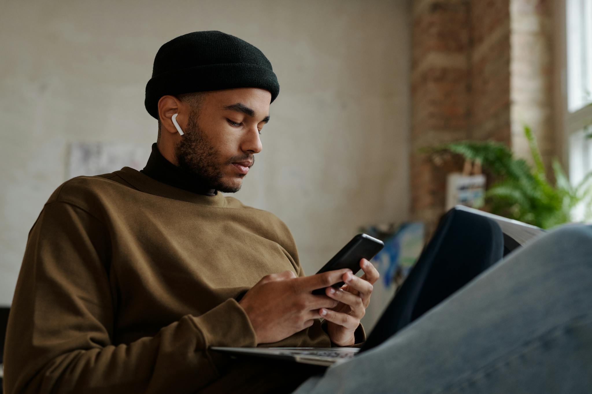 Young man with wireless earbuds using smartphone indoors, casual style in a relaxed setting.